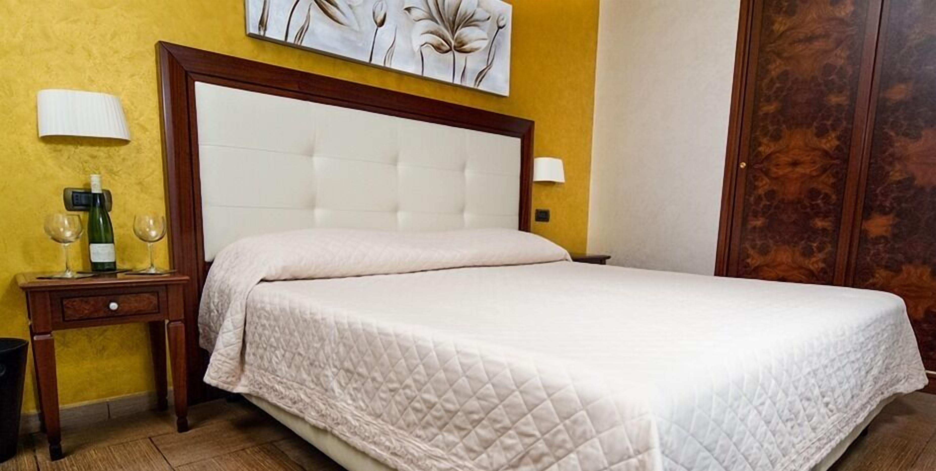 Une chambre d’hôtel avec un grand lit, une table de chevet avec une bouteille et des verres, et une lampe fixée au mur.