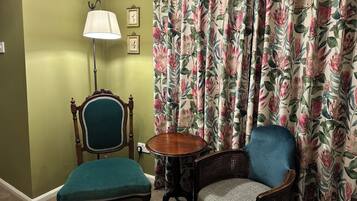 A room with a green wall, a wooden chair with a green cushion, a wicker chair with a patterned cushion, a small wooden table, and floral curtains.