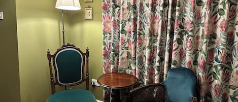 Um quarto com parede verde, uma cadeira de madeira com almofada verde, uma cadeira de vime com almofada estampada, uma pequena mesa de madeira e cortinas florais.