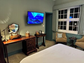 A hotel room with a bed, a desk with a lamp, a television mounted on the wall, a window with curtains, and a small table with two bottles.