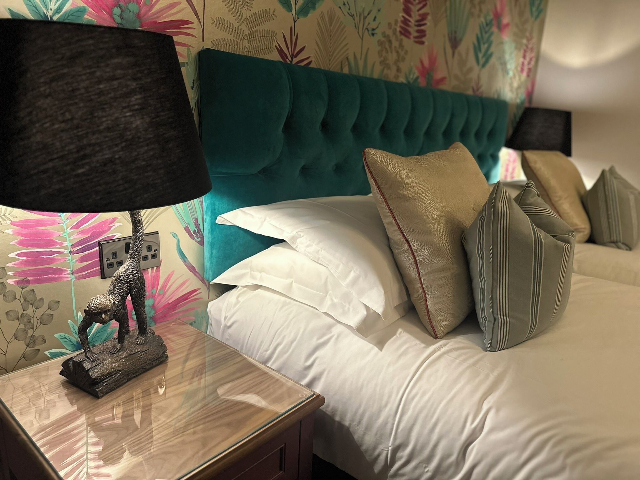 A neatly made bed with white and striped pillows, a teal headboard, and a small table with a lamp and a sculpture.