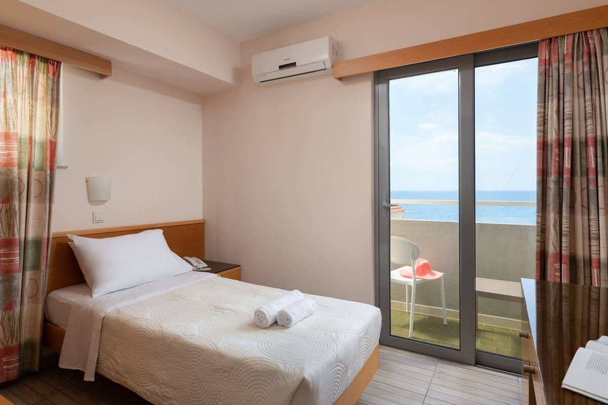 A hotel room with a bed, a bedside table, a phone, an air conditioning unit, a window with a view of the sea, and a balcony with a chair.