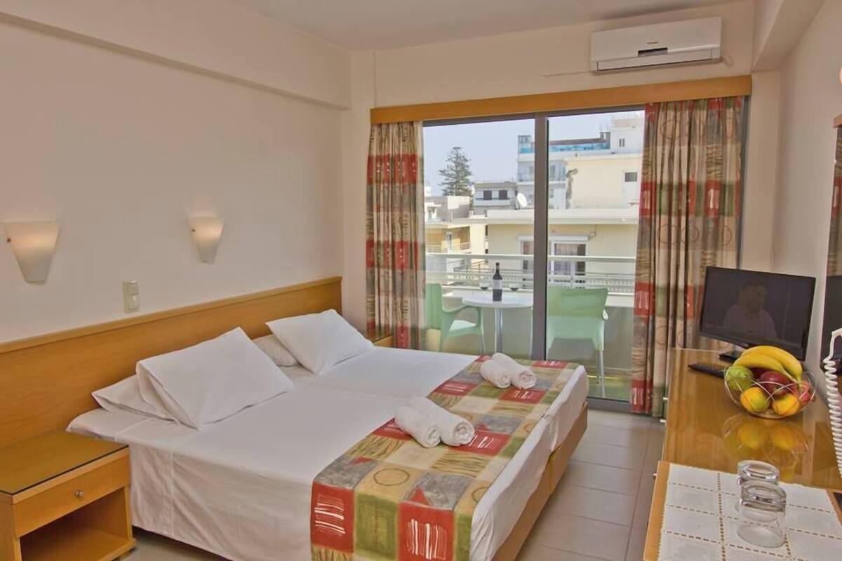 A hotel room with a bed, a desk with a TV, a window with curtains, and a balcony with a table and chairs.