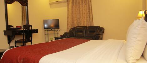 A hotel room with a large bed, a television, a leather chair, a desk with a mirror, and an air conditioning unit.