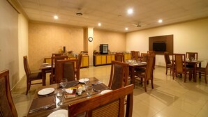 Un coin repas avec des tables et des chaises en bois, un espace cuisine équipé d’une télévision et une horloge au mur.