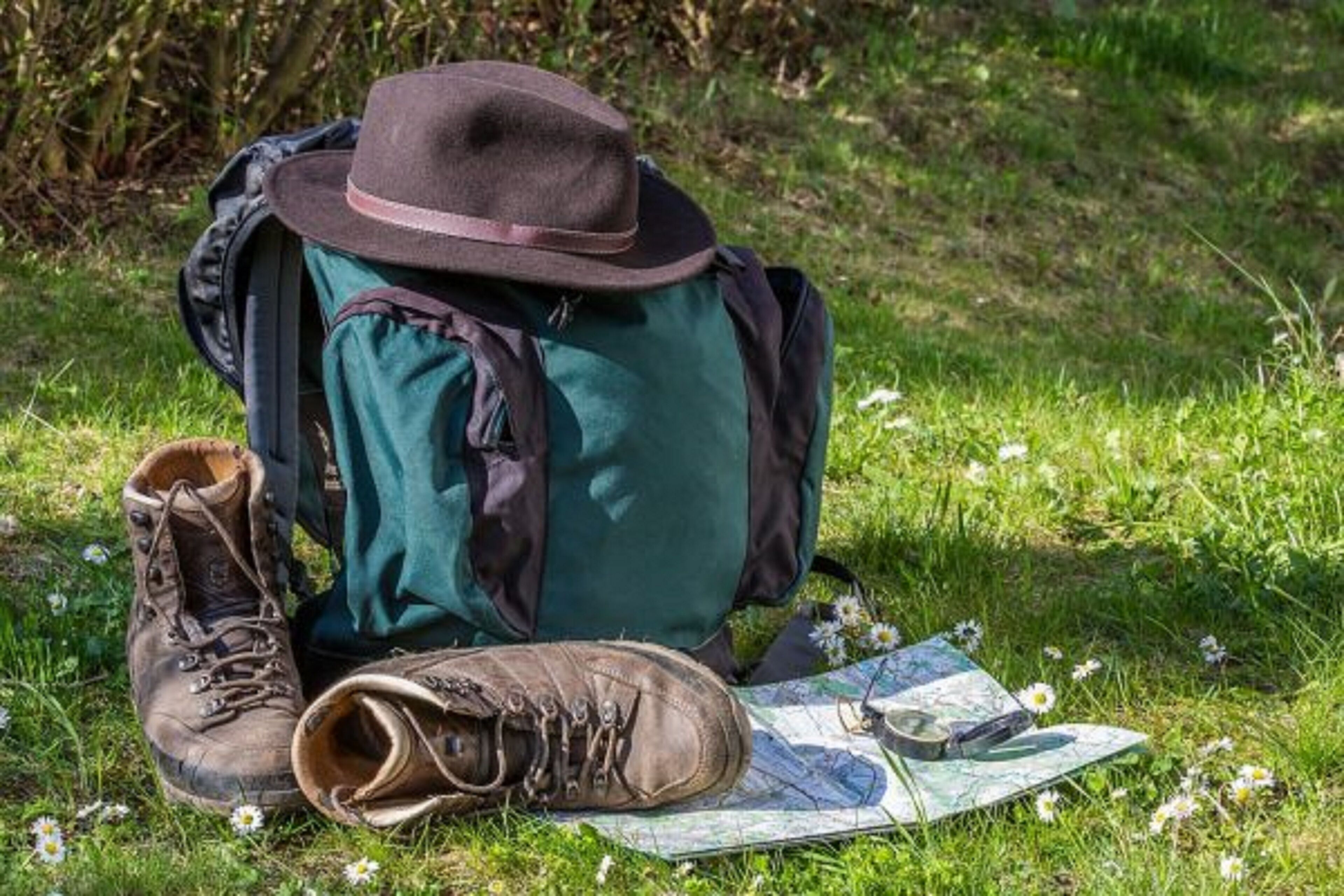 Una mochila verde, un sombrero marrón y un par de botas de senderismo marrones sobre hierba, junto con un mapa y una brújula.