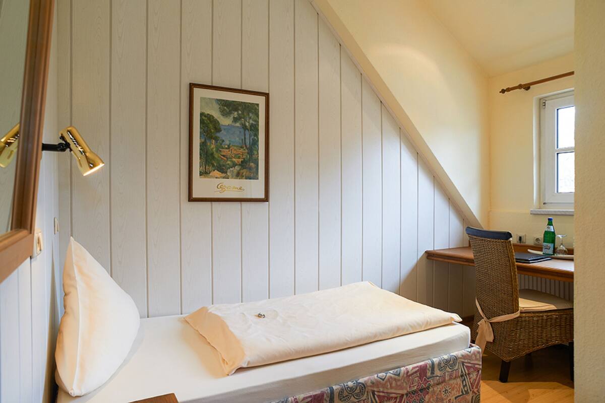 A bedroom with a bed, a chair, a desk, a window, and a painting on the wall.
