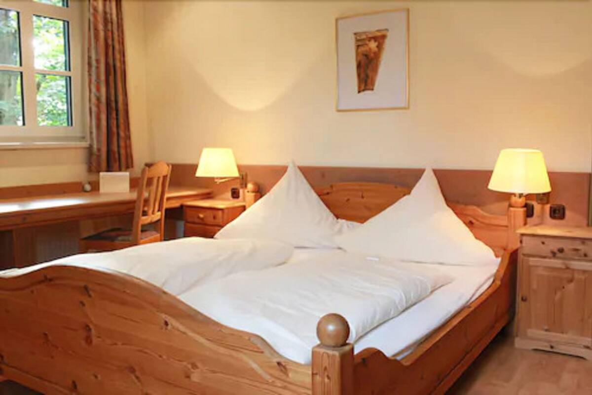 A wooden bed with white linens, two bedside lamps, a chair, a desk, and a framed picture on the wall.