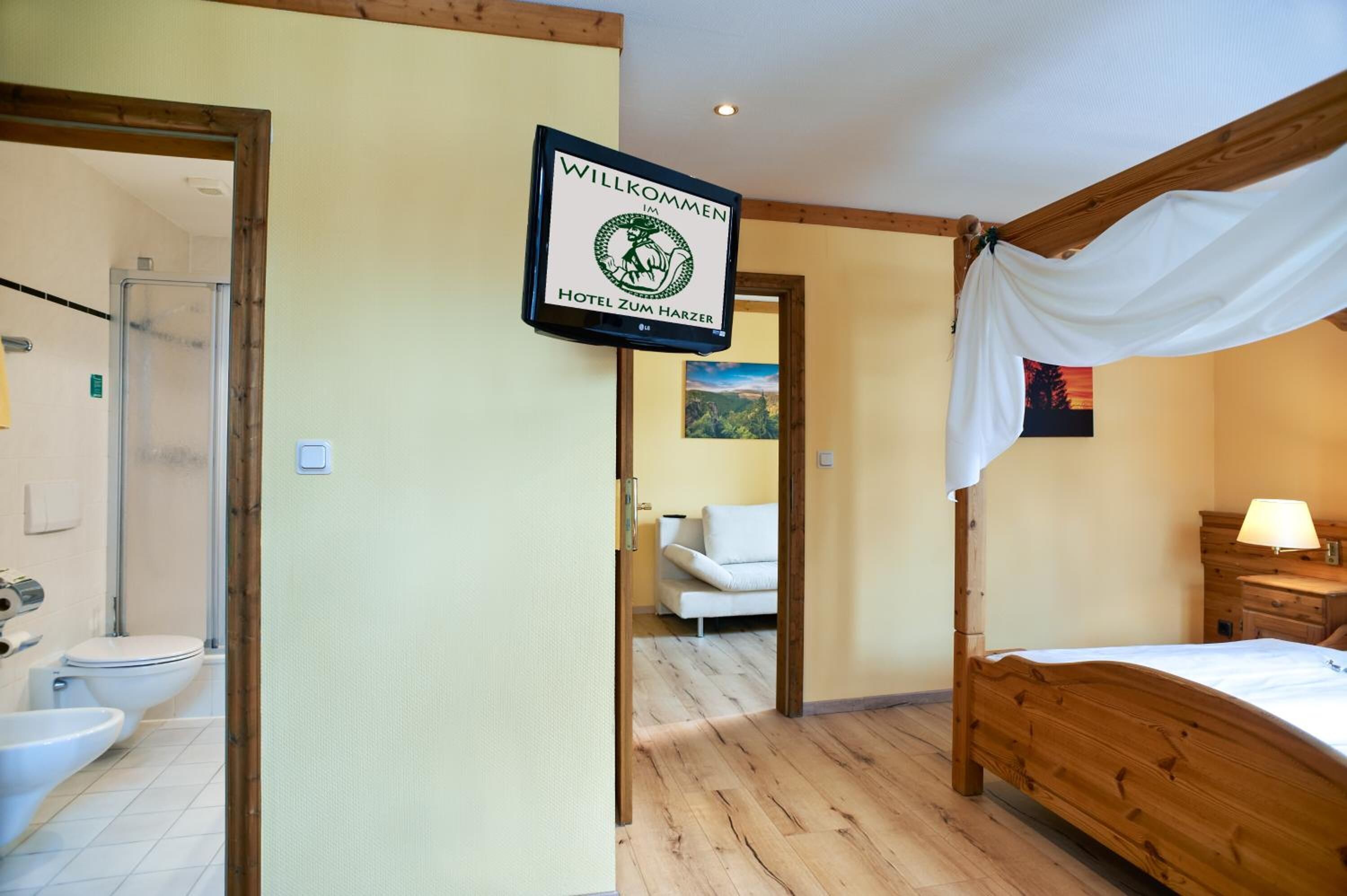 A hotel room with a wooden bed, a white canopy, a bathroom with a shower, and a sign that reads "WILLKOMMEN HOTEL ZUM HARZER".