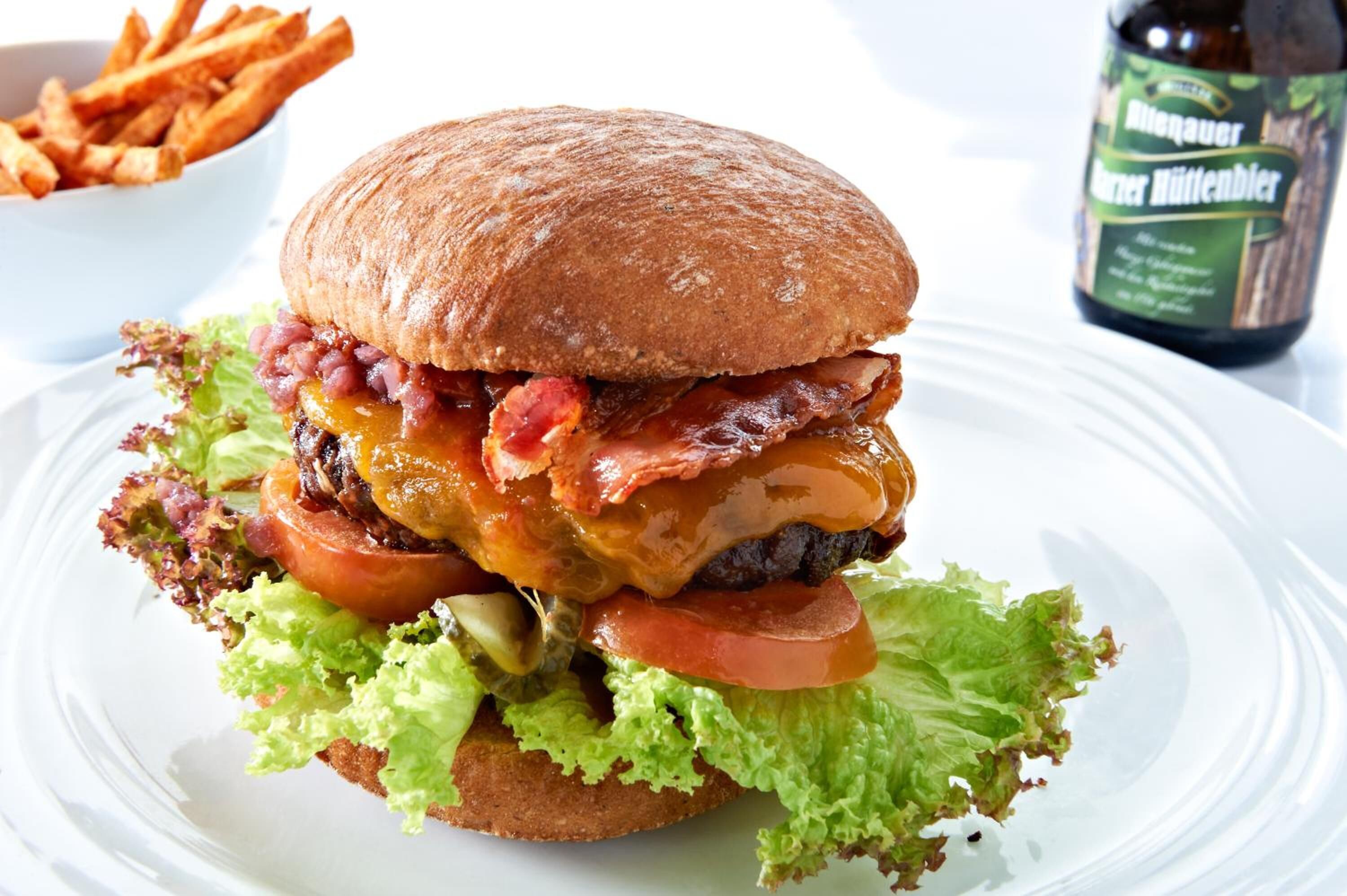 A burger with lettuce, tomato, cheese, and bacon on a bun, accompanied by fries and a bottle of beer.