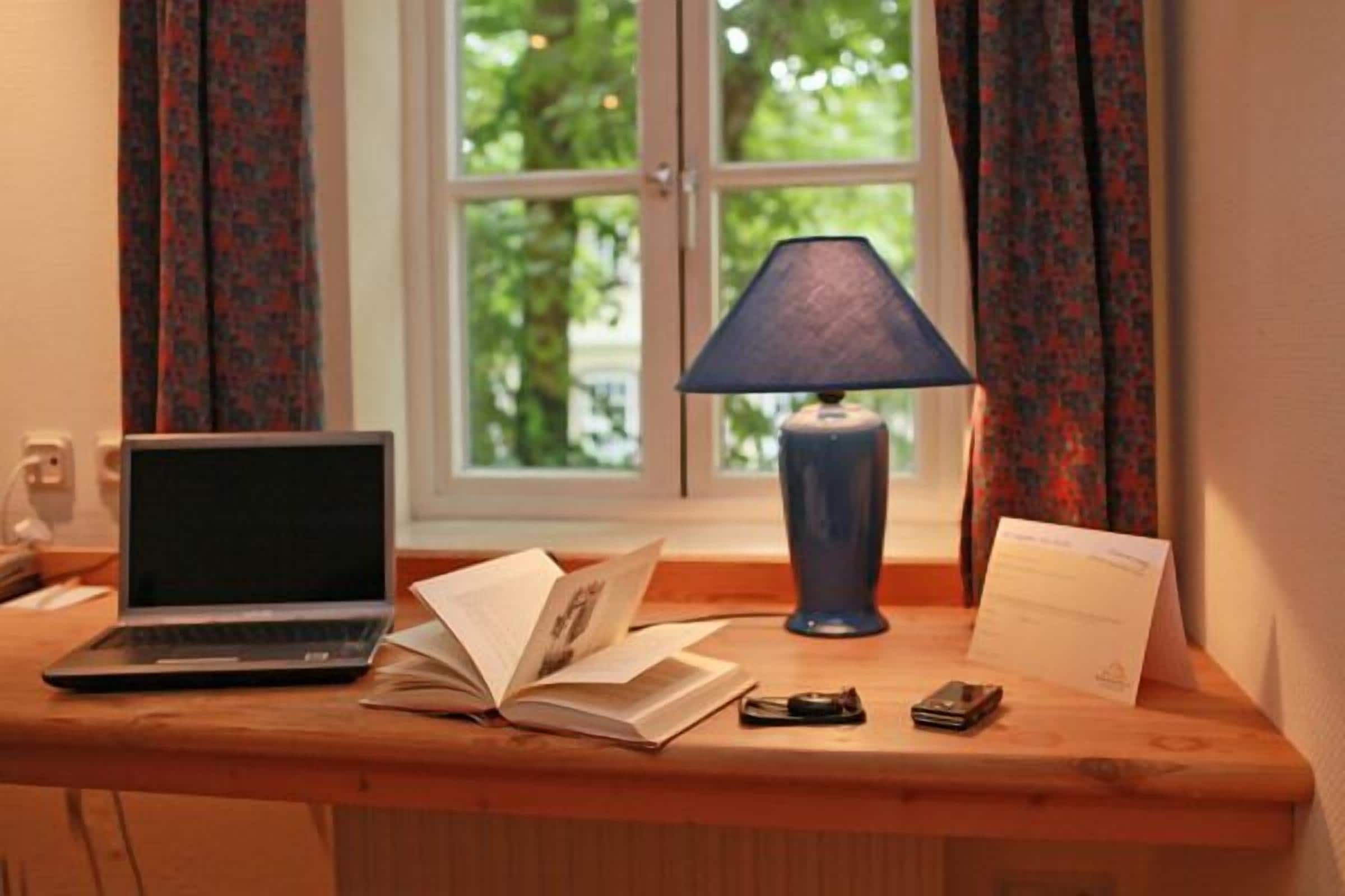 A wooden desk with a laptop, an open book, a lamp, a pair of glasses, and a mobile phone.