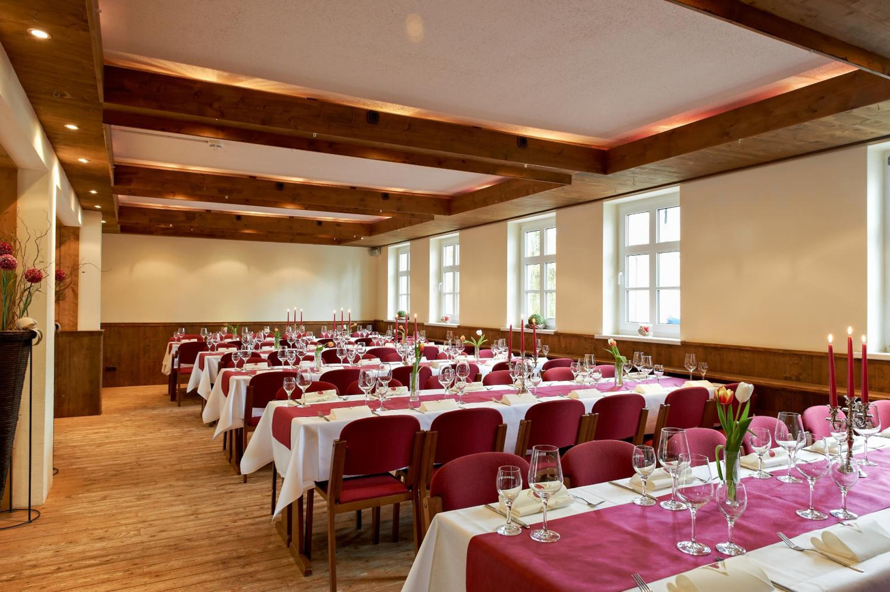 Uma sala de banquetes com longas mesas preparadas para refeições, vigas de madeira no teto e grandes janelas.