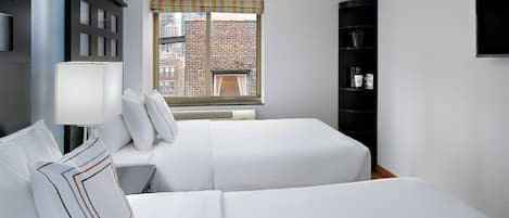 Habitación de hotel con dos camas, una ventana que da a edificios, una estantería negra y un televisor montado en la pared.