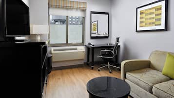 Habitación de hotel con televisor de pantalla plana, escritorio con silla, sofá con cojín verde, mesa redonda negra y ventana grande con vista a edificios.