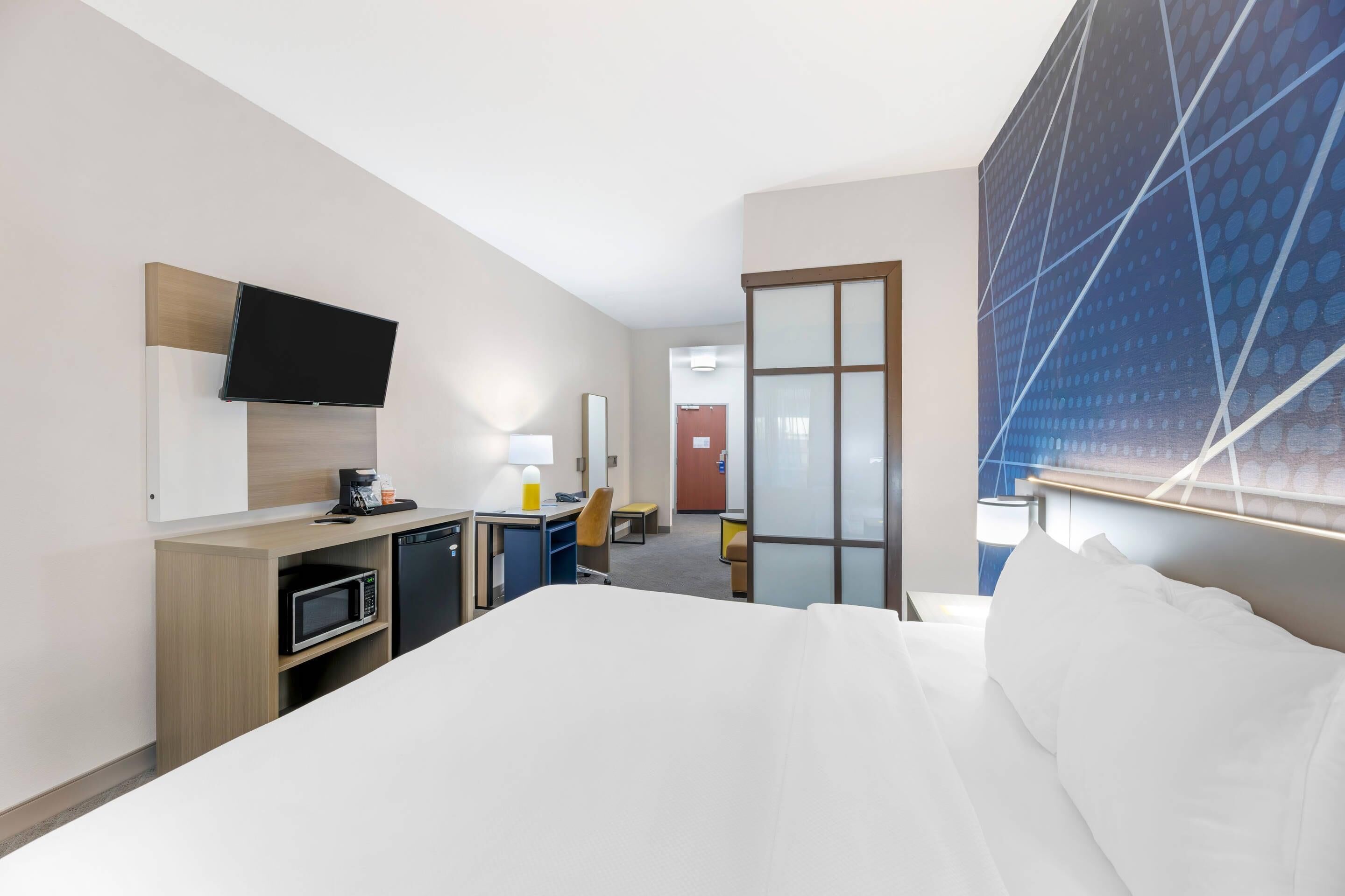 A hotel room with a bed, a desk with a microwave, a chair, a TV mounted on the wall, and a large window with a view of the cityscape.