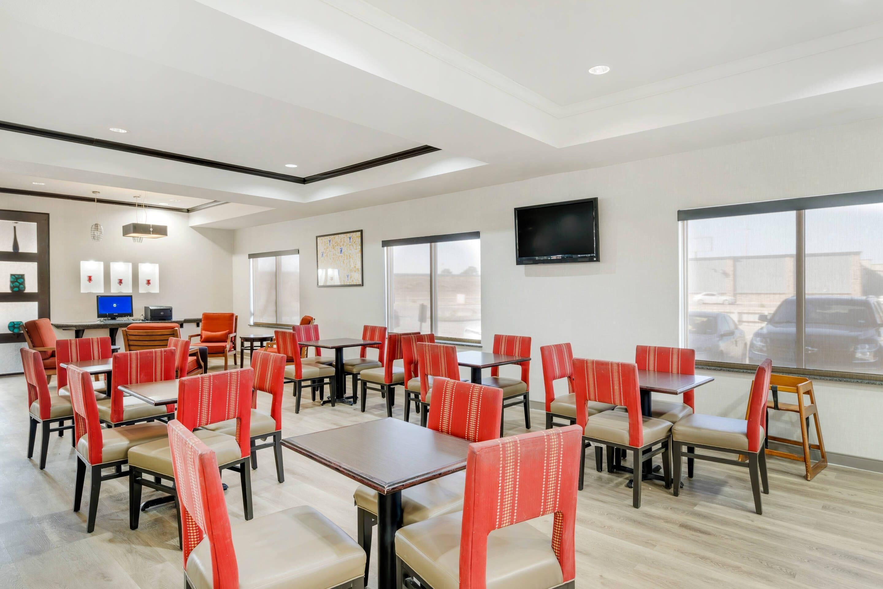 A modern dining area with multiple tables and chairs, a flat-screen TV mounted on the wall, and large windows offering a view of the parking lot.