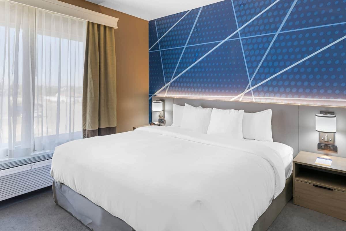 A hotel room with a large bed, a nightstand, a wall-mounted lamp, and a window with sheer curtains.