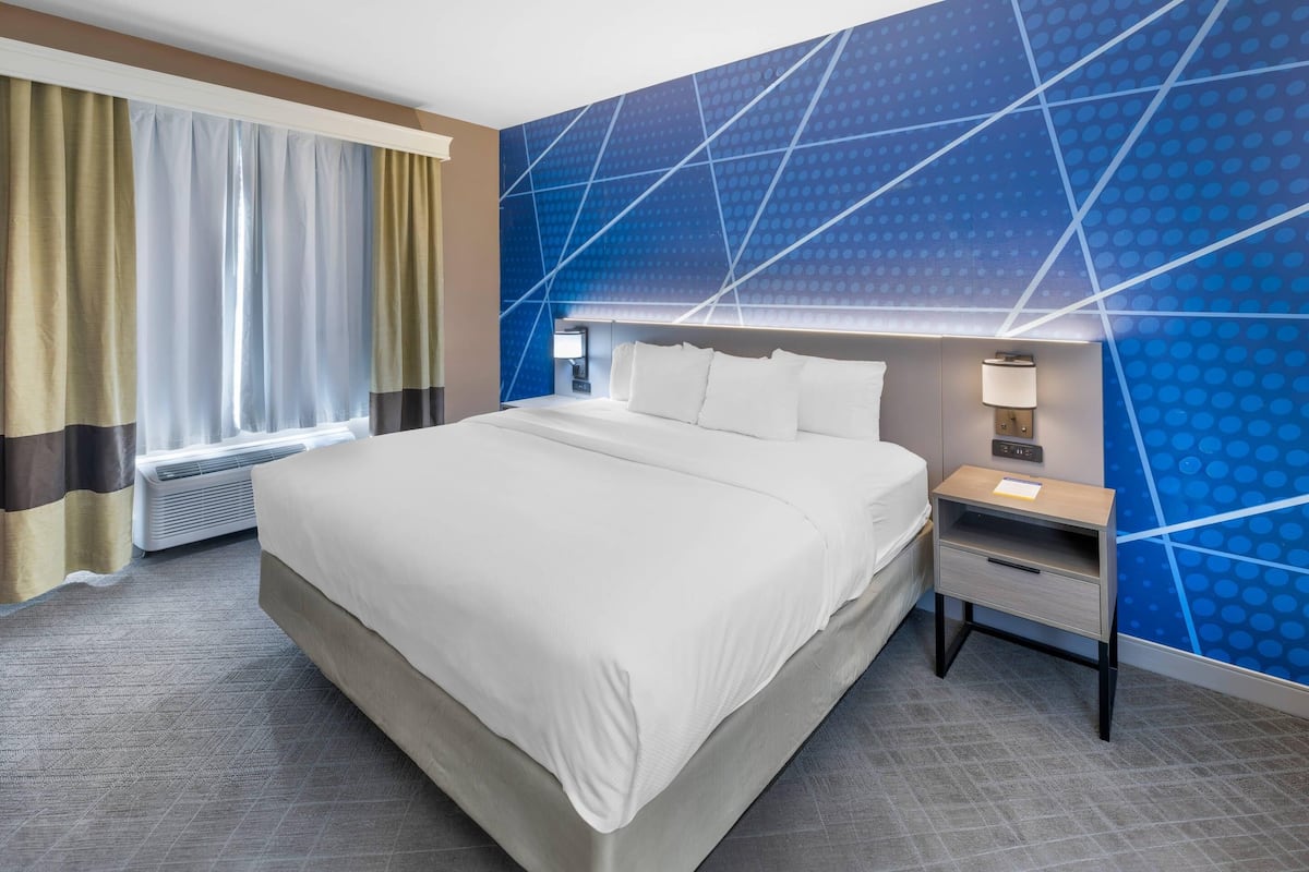 A hotel room with a large bed, a nightstand with a lamp, and a wall with a geometric pattern.