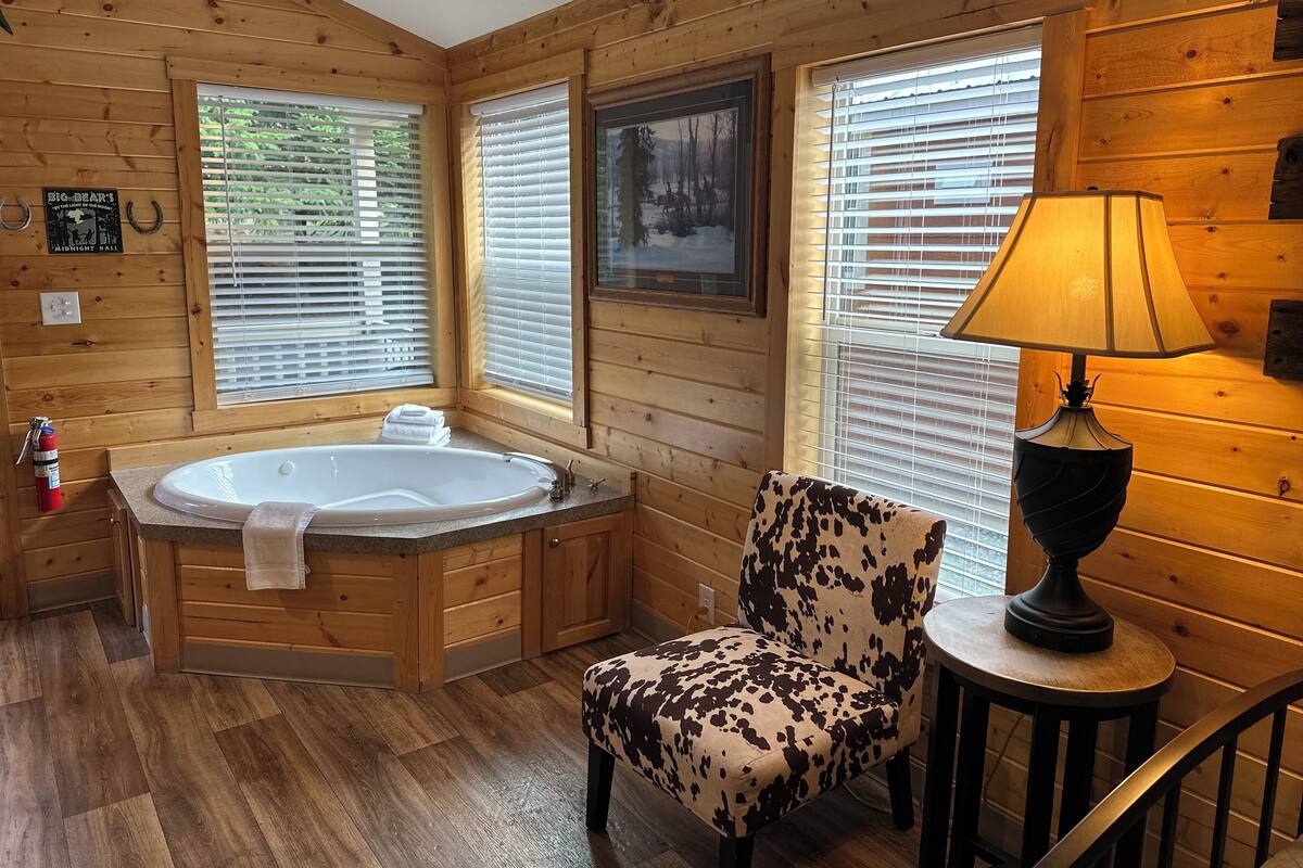 Ein Badezimmer einer Holzhütte mit einer großen Badewanne, einem Sitzmöbel mit Kuhmuster und einem Tisch mit einer Lampe.
