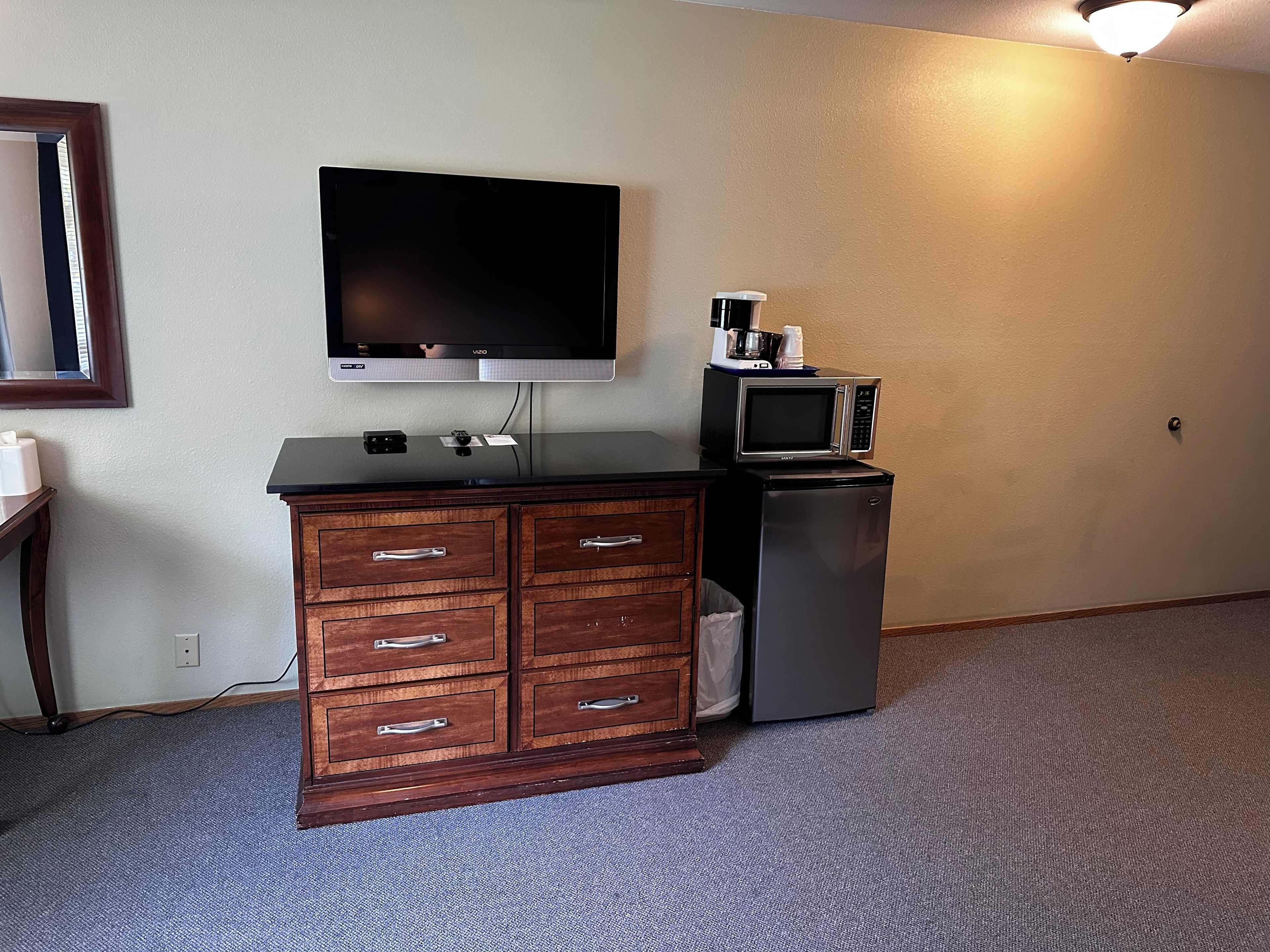 Habitación con televisión de pantalla plana montada en la pared, cómoda de madera con cajones, microondas y refrigerador pequeño.