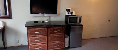 A room with a flat-screen TV mounted on the wall, a wooden dresser with drawers, a microwave, and a small refrigerator.