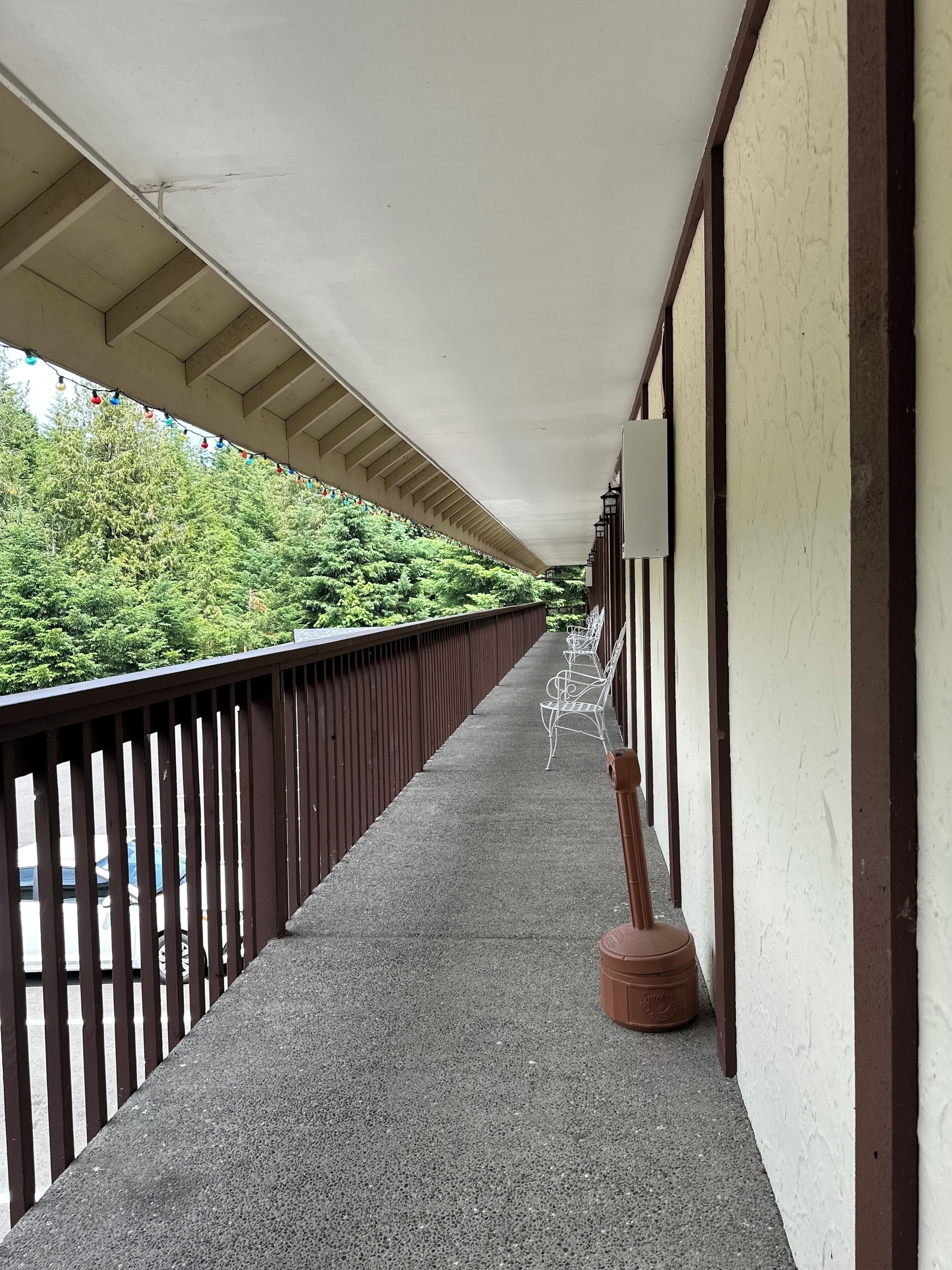A covered corridor with a concrete floor, metal railings, and a row of white chairs.