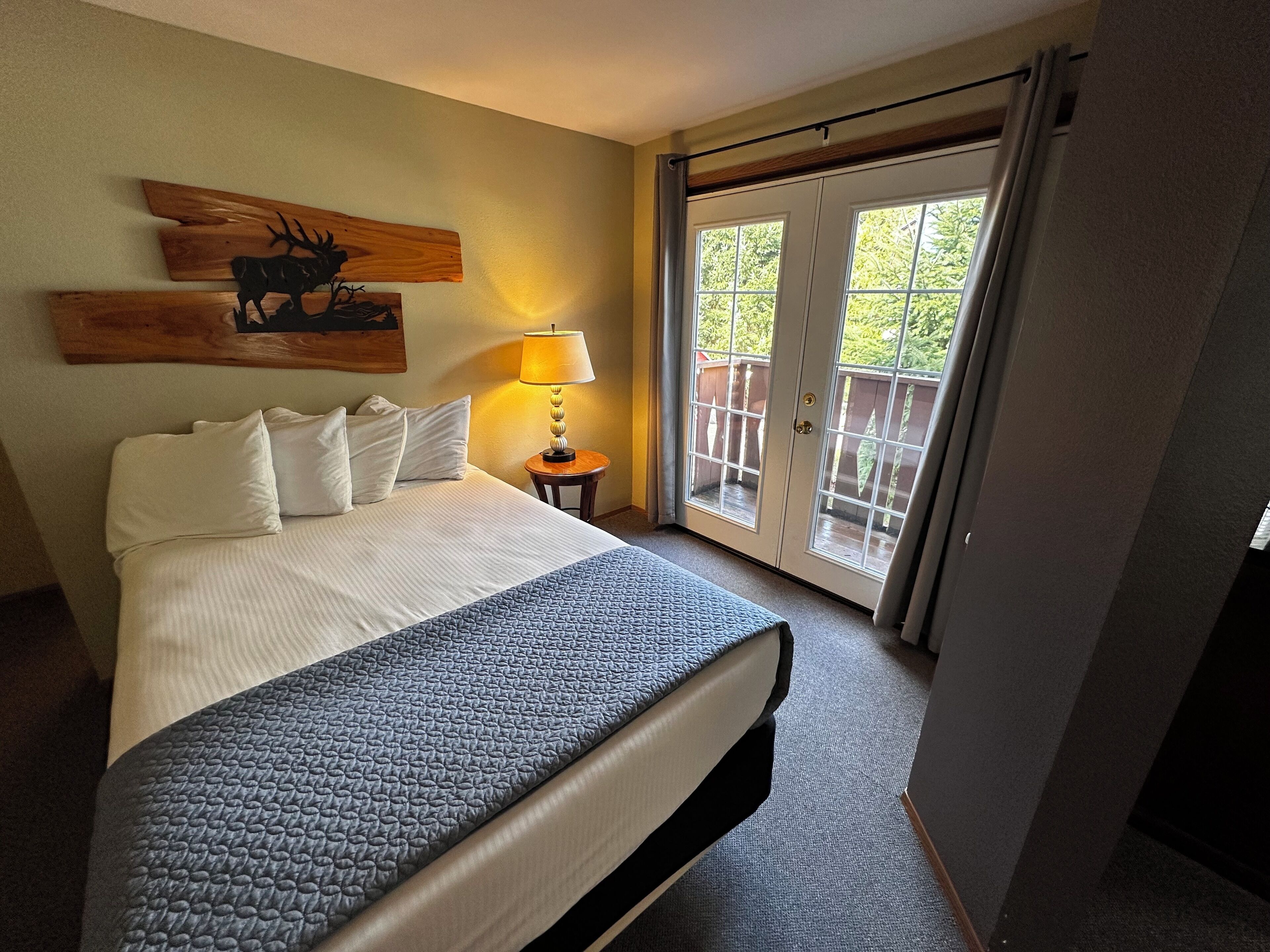 Habitación de hotel con cama, mesita de noche con lámpara, un adorno de pared de madera y una puerta corrediza de cristal que da a un espacio exterior.