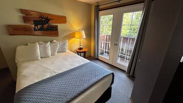 A hotel room with a bed, a nightstand with a lamp, a wooden wall art piece, and a sliding glass door leading to an outdoor area.