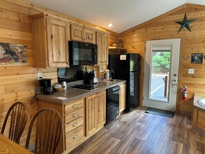 A wooden cabin-style kitchen with wooden cabinets, a black microwave, a black refrigerator, and a dining table with chairs.