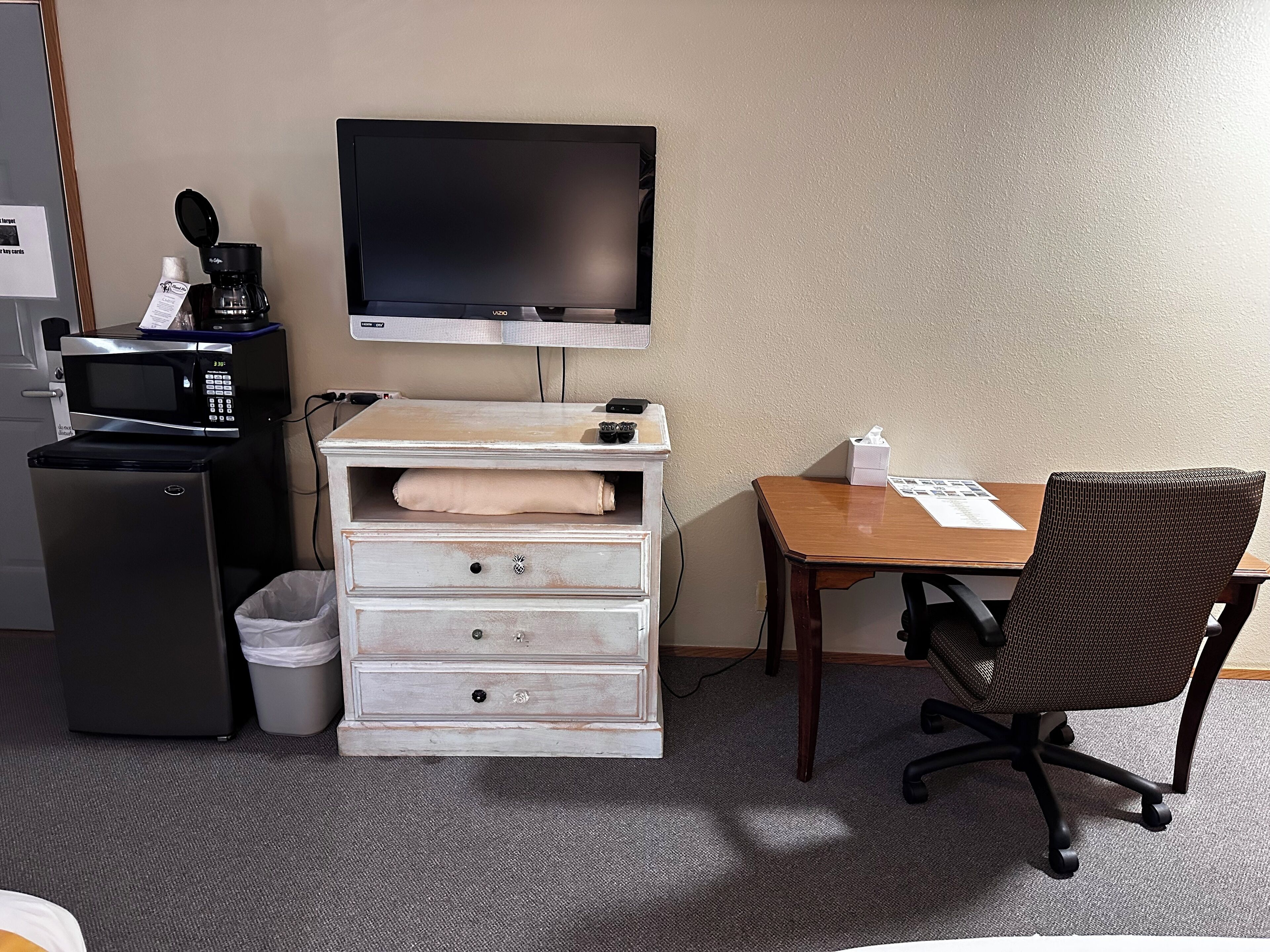 A compact hotel room with a kitchenette, a flat-screen TV, a wooden desk, and a wicker chair.