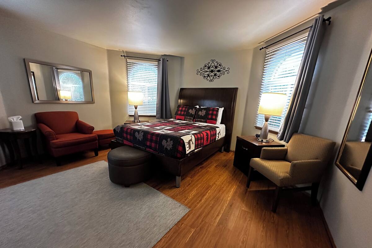 Ein Schlafzimmer mit einem Bett, zwei Stühlen, einem kleinen Tisch, einem Spiegel und einem Fenster mit Jalousien.