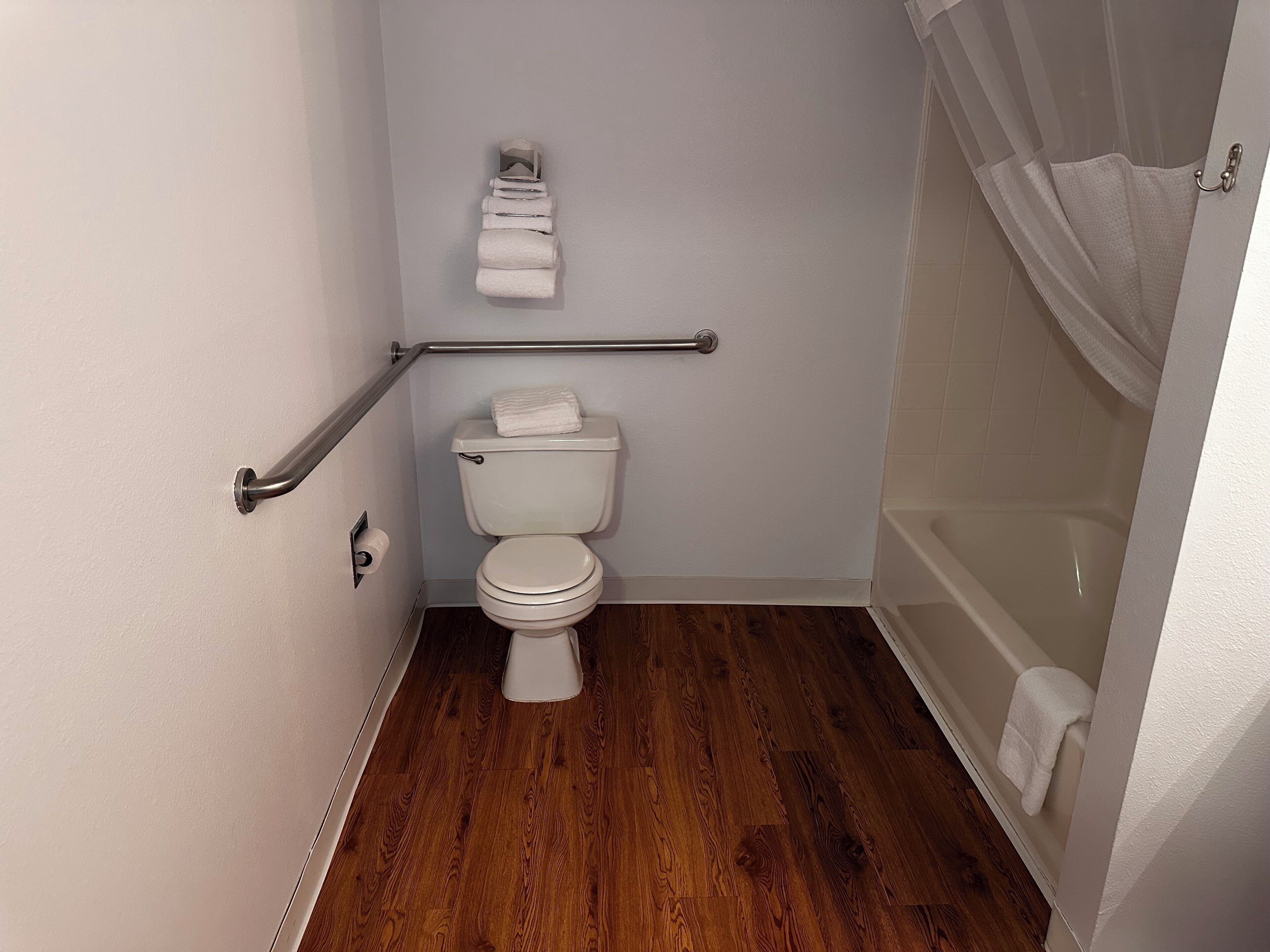Une petite salle de bain propre, équipée de toilettes, d’une barre d’appui et d’un rideau de douche.