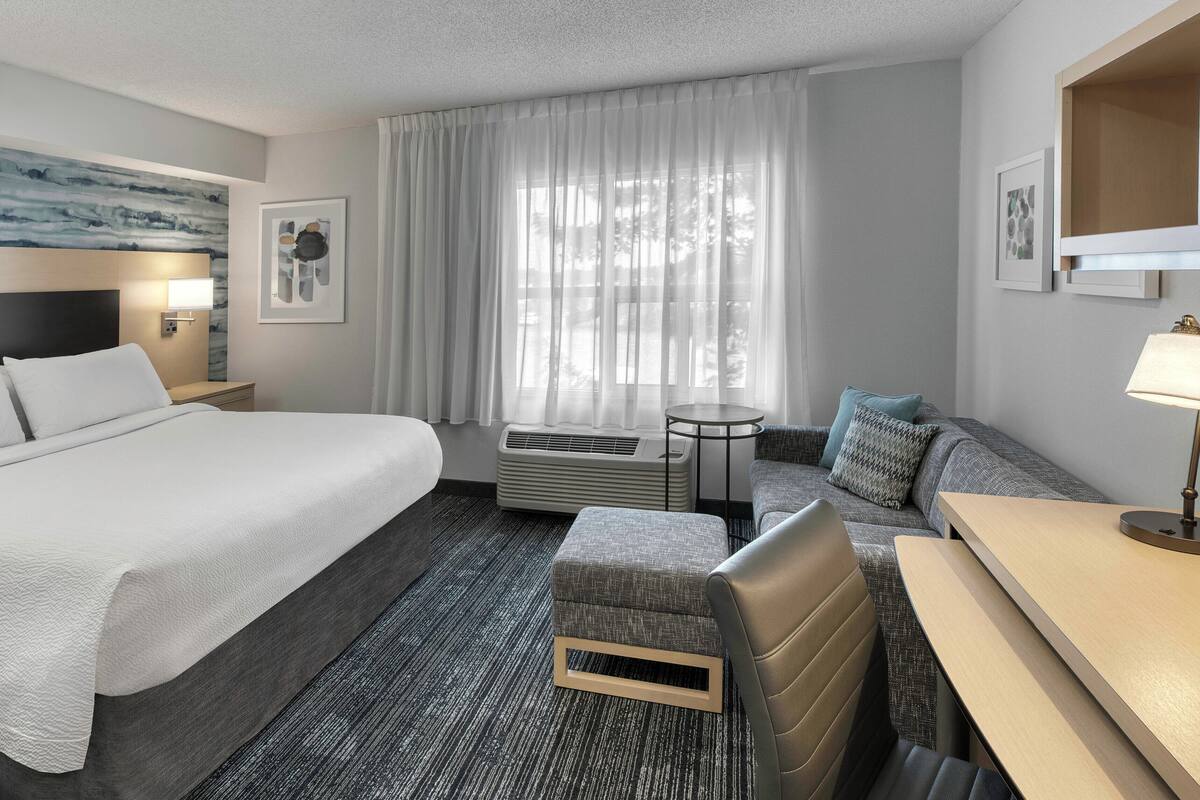 Une chambre d’hôtel avec un grand lit, un canapé, une petite table et un bureau. Il y a une fenêtre avec des rideaux, un climatiseur fixé au mur et une lampe sur le bureau.