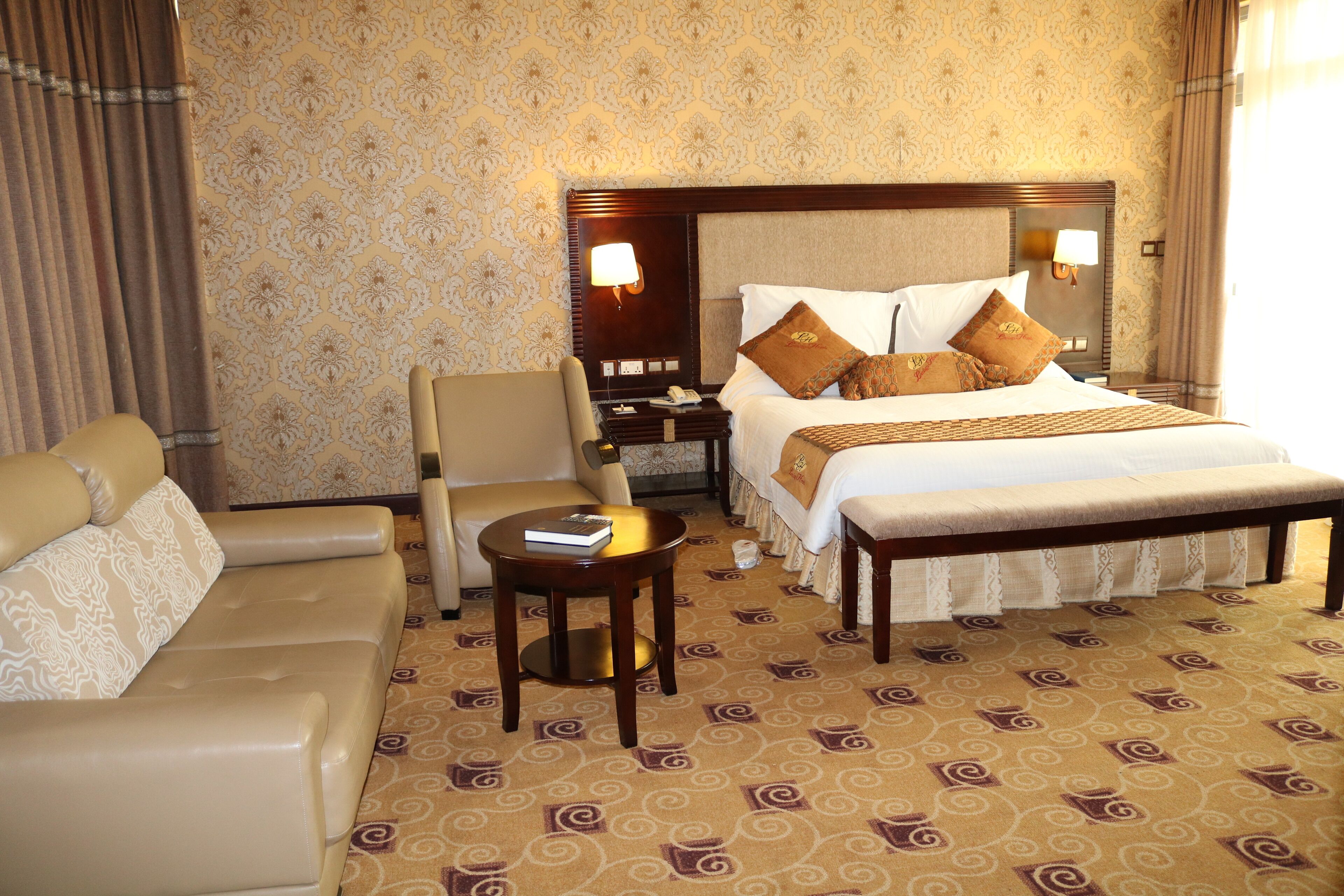 A hotel room with a bed, a sofa, a chair, a small table, and a nightstand with a lamp.
