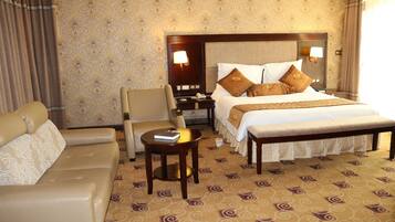 A hotel room with a bed, a sofa, a chair, a small table, and a nightstand with a lamp.