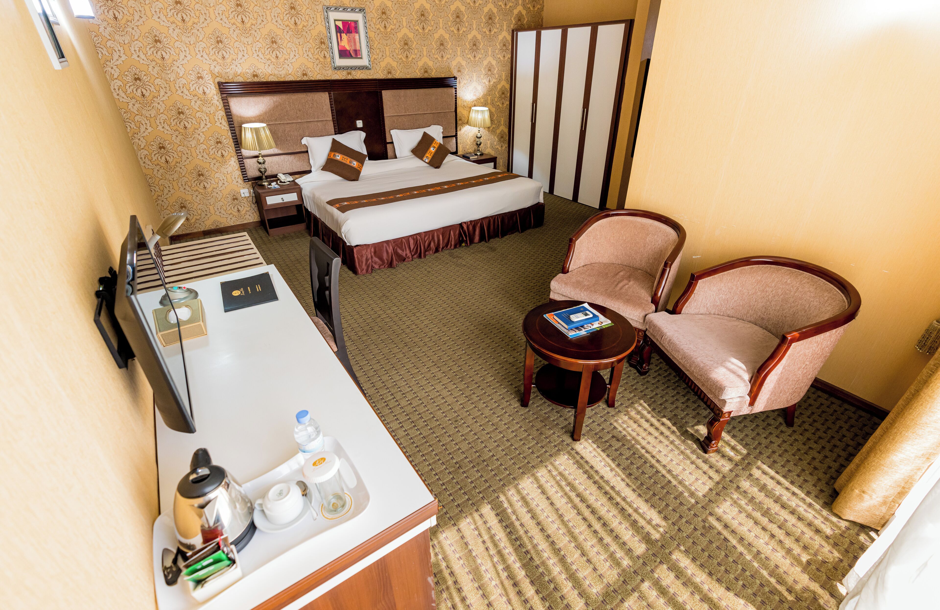 A hotel room with a bed, bedside table, chair, and a desk with a kettle, cups, and a book.