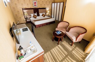 Quarto de hotel com cama, mesa de cabeceira, cadeira e escrivaninha com chaleira, xícaras e um livro.