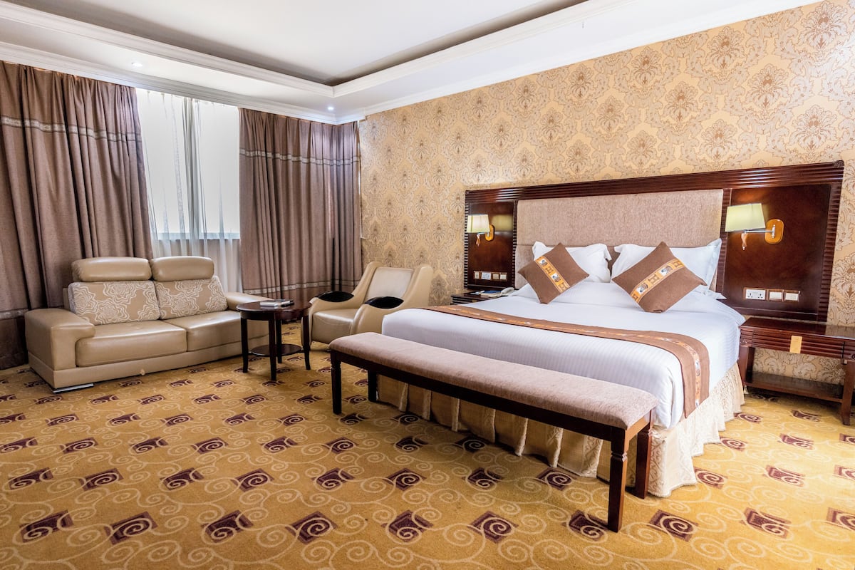 A hotel room with a large bed, a sofa, a chair, a small table, and a bench.
