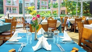 A dining area with tables set for two, featuring a QR code for payment, a vase with flowers, and a view of a building with balconies.