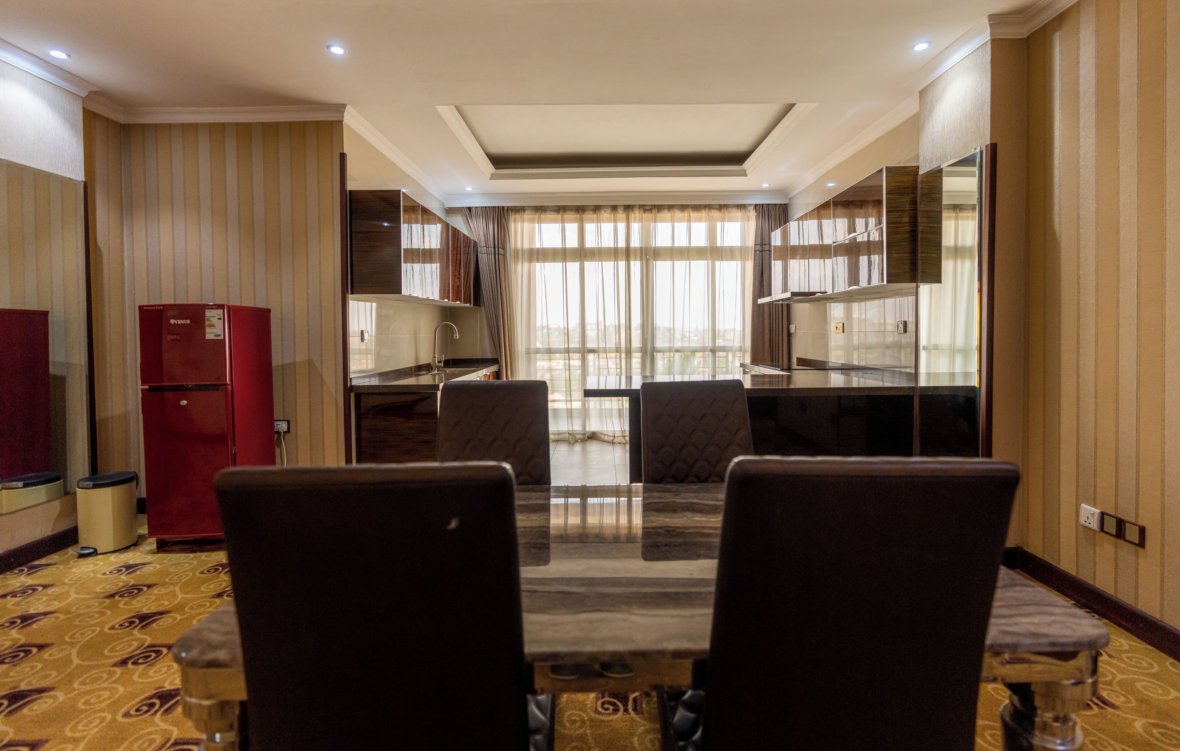 A hotel room with a dining area featuring a table and chairs, a red refrigerator, and a kitchen area with a sink and cabinets.