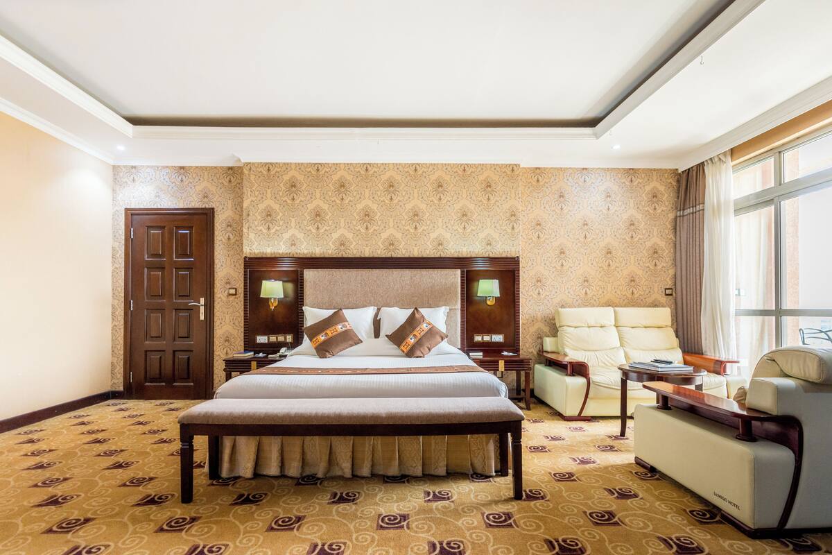 A hotel room with a large bed, a sitting area with a sofa and armchairs, a wooden headboard, and a patterned carpet.