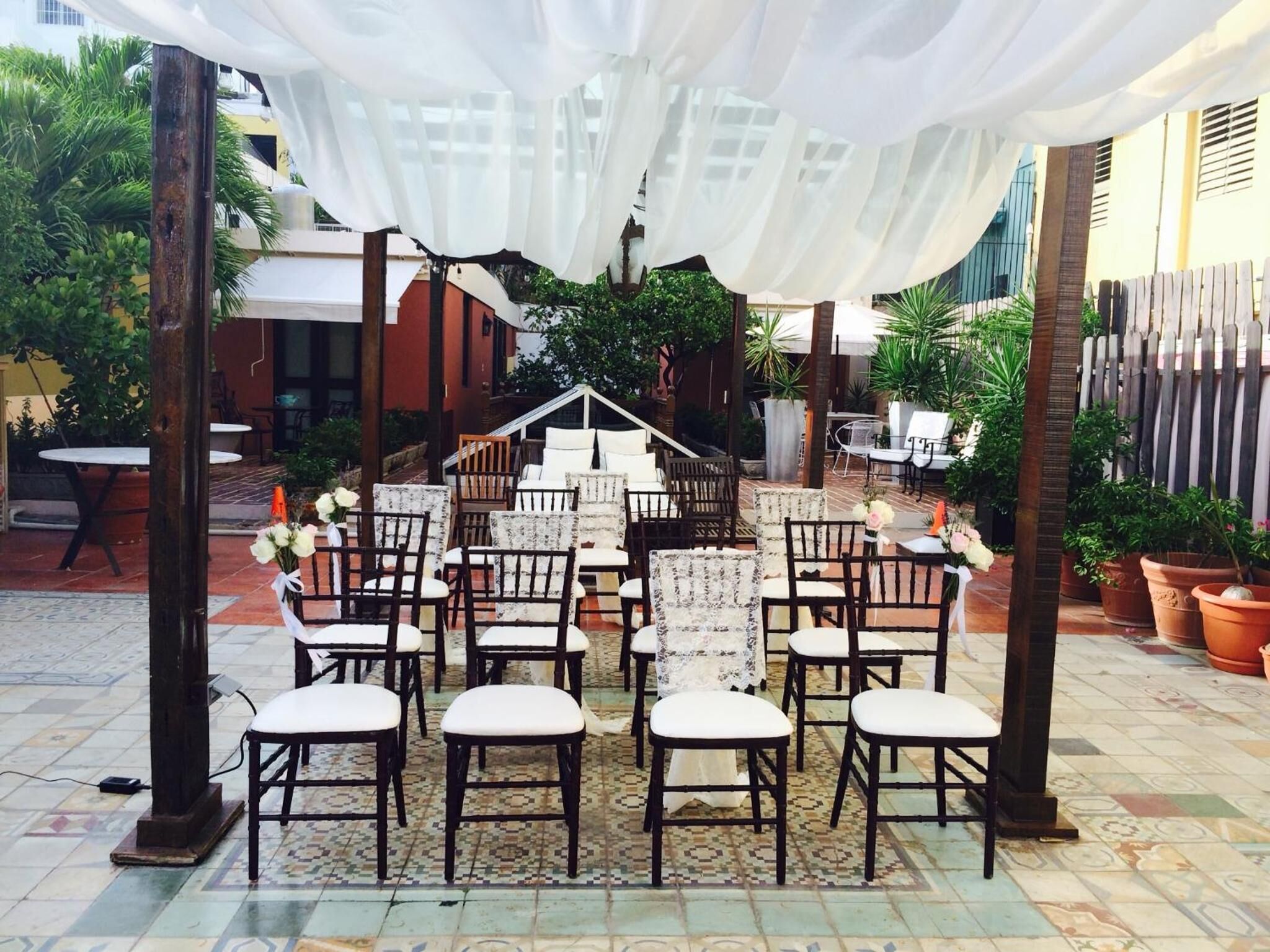 An outdoor patio with rows of chairs and tables, surrounded by potted plants and a white canopy.