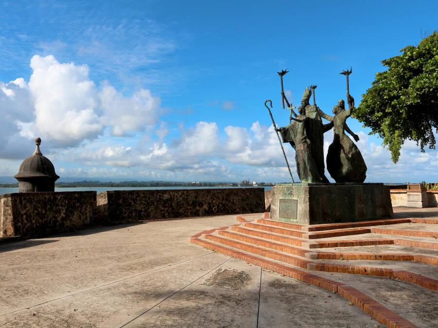 Una estatua de tres figuras sobre un pedestal con escalones que llevan hacia ella, dominando un cuerpo de agua.