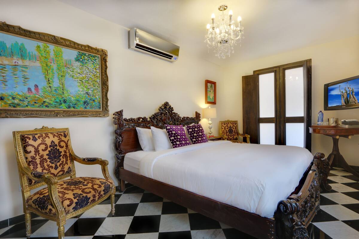 Een hotelkamer met een zwart-wit geruit vloerkleed, een groot bed met een decoratief hoofdeinde, een stoel met een bekleedde zitting en een schilderij aan de muur.