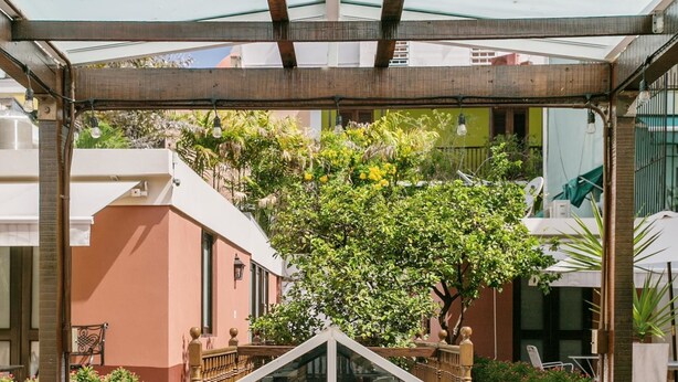 Een bed met witte lakens en twee hoeden, een balkon met glazen wanden met uitzicht op groen en gebouwen, en een houten pergola.