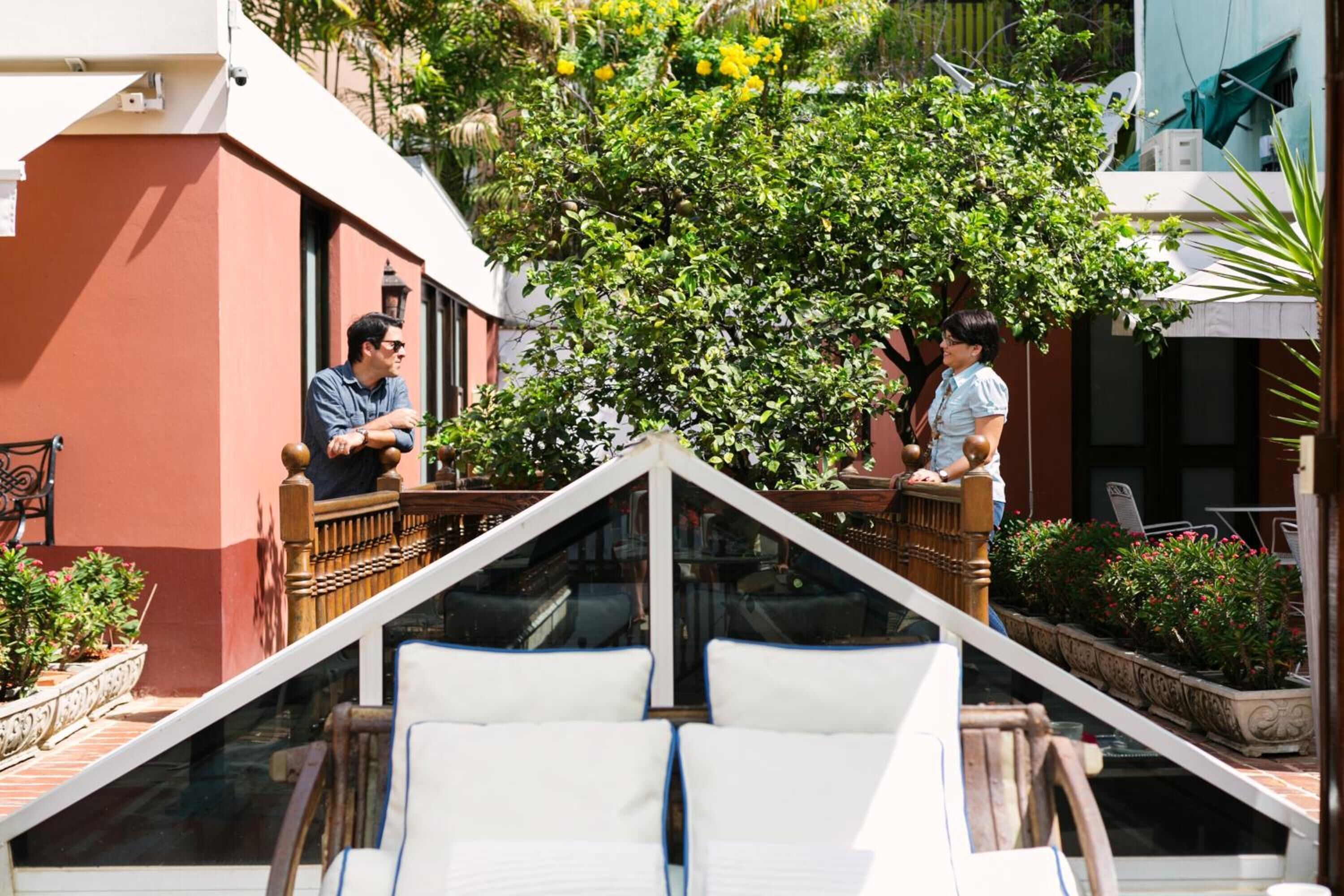Ein Balkon mit zwei sitzenden Personen, einem Blumentopf und Blick auf ein Gebäude.