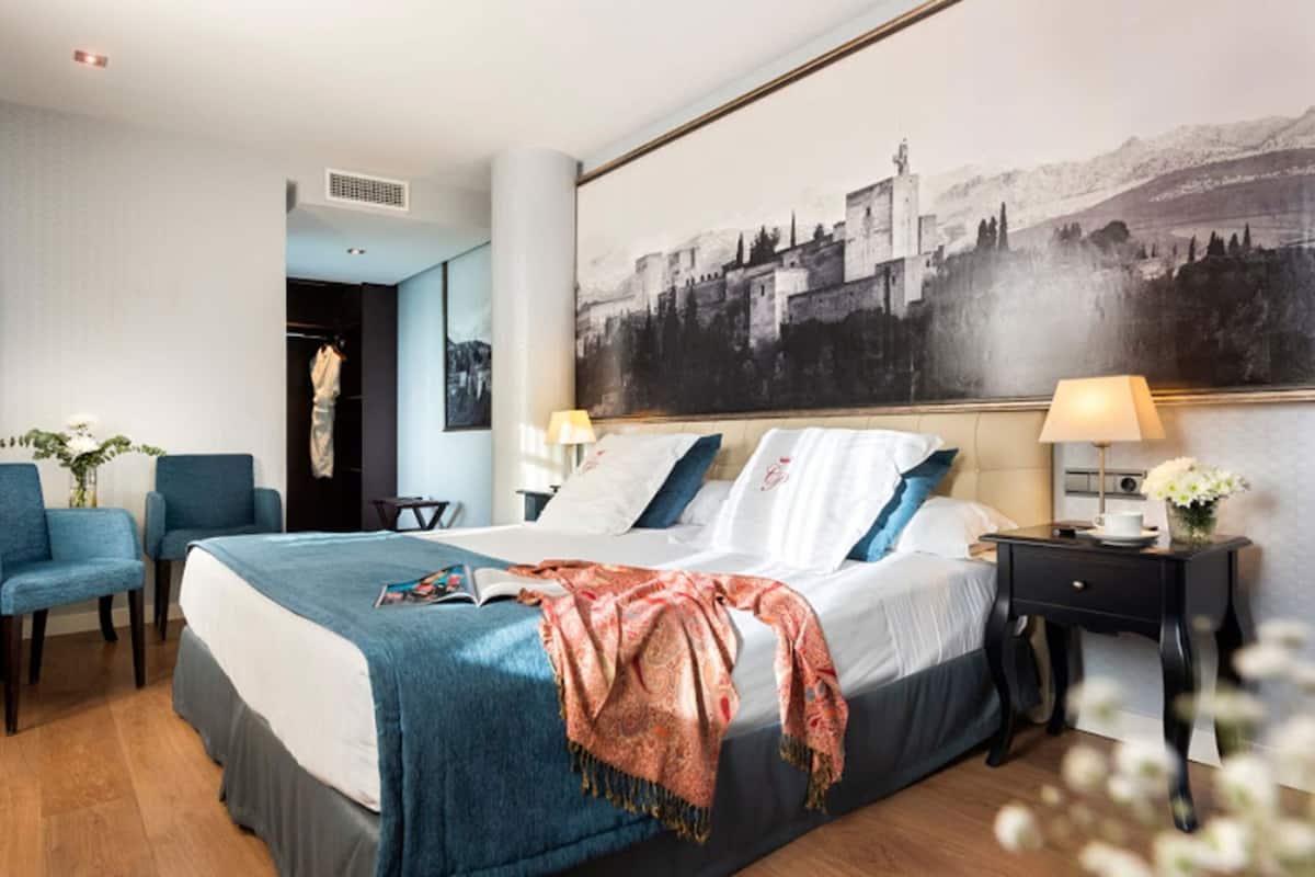 Habitación de hotel con cama, dos sillas, una mesita de noche, lámpara, un cuadro y una puerta que da a otra habitación.