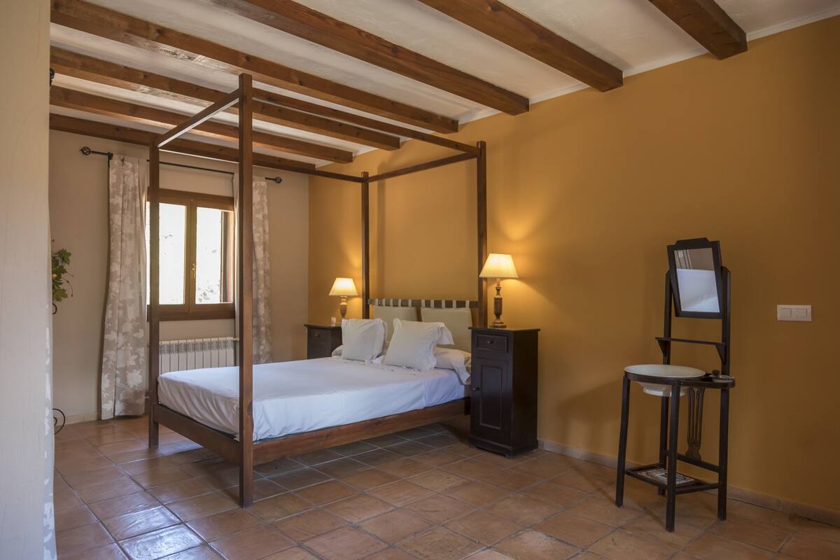 Un dormitori amb un llit amb cortines, dues taules de nit amb làmpades, una taula rodona petita i un mirall a la paret.