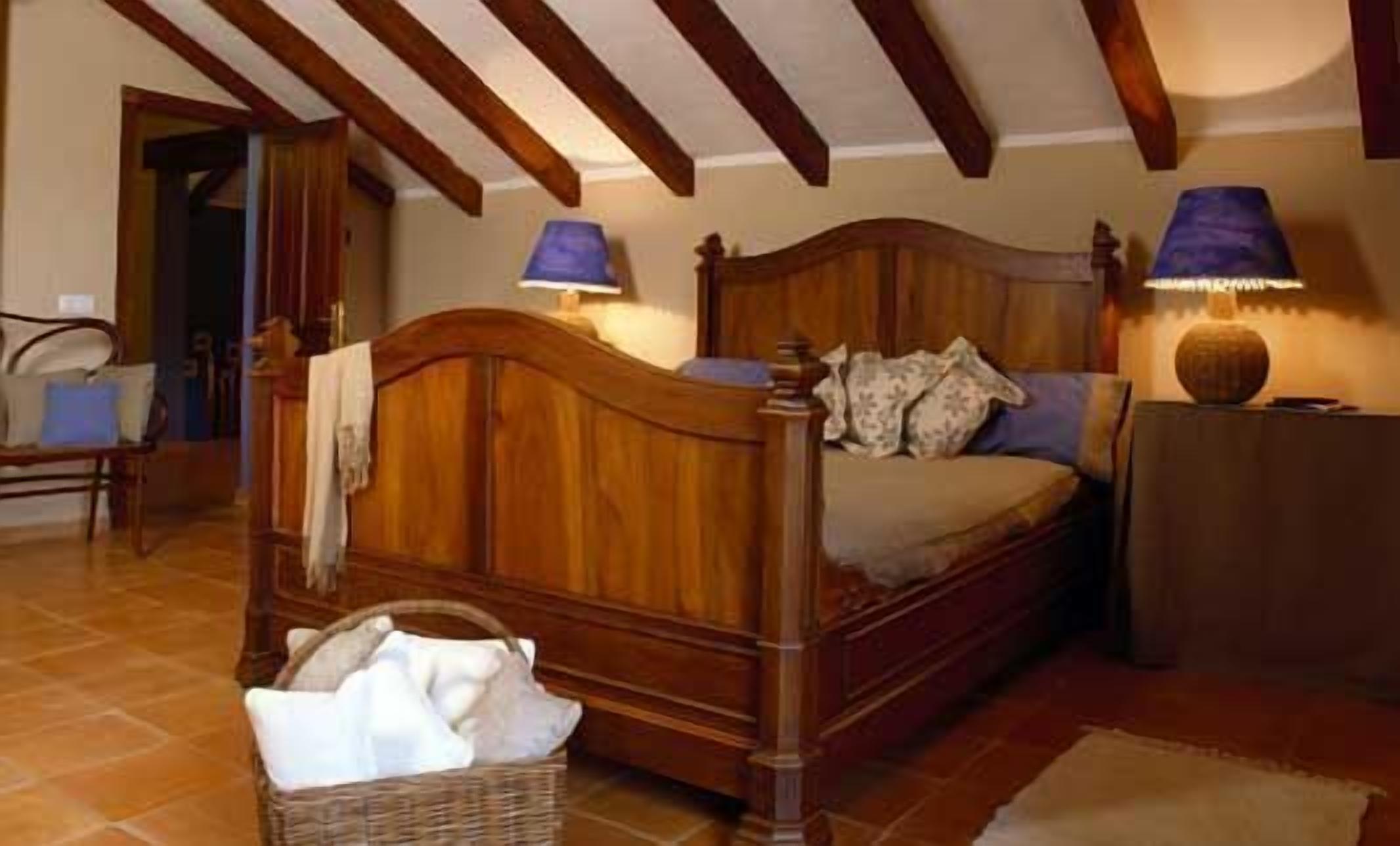 Un dormitori amb llit de fusta, dues làmpades, una tauleta de nit, una cistella de vimet i una cadira.