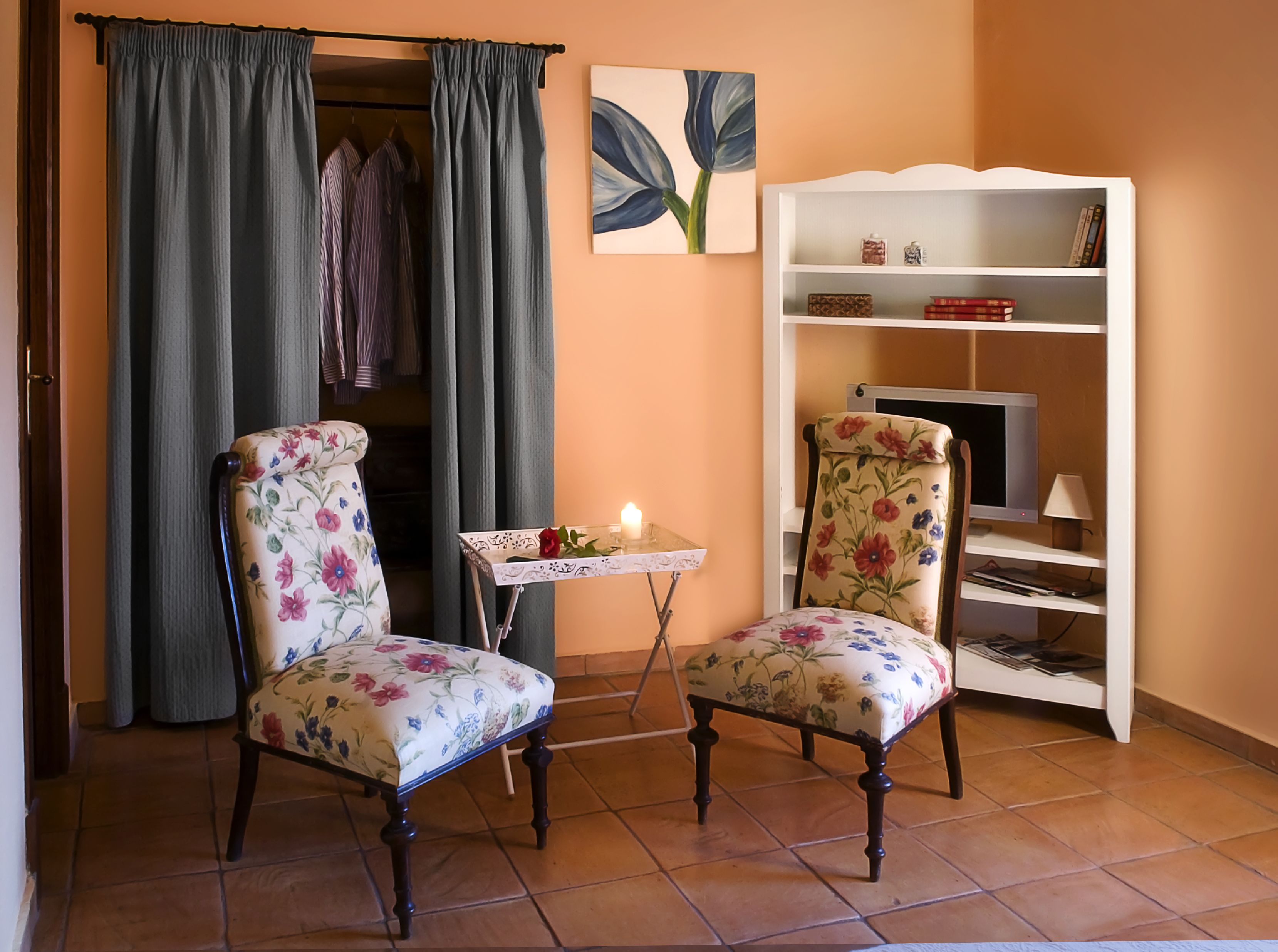 Een kamer met twee armstoelen met bloemenprint, een kleine tafel met een kaars en een boekenkast tegen een muur.